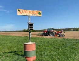 "Rent an Acker" Holzschild im Vordergrund, im Hintergrund ein roter Traktor vor einem blauen Himmel