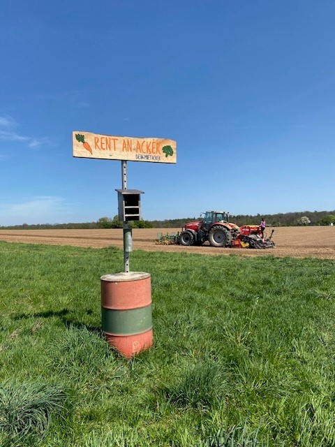 "Rent an Acker" Holzschild im Vordergrund, im Hintergrund ein roter Traktor vor einem blauen Himmel